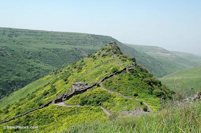 A green peak with stone paths and structures leading up the peak, rolling hills in the background