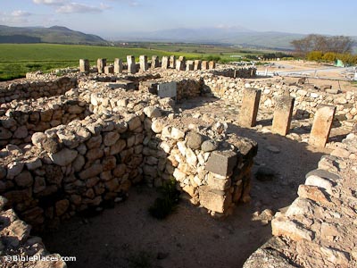 A maze of stone walls and pillars forming a roofless structure with green fields in the background