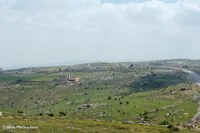 A green hill with scattered trees, bushes, rocks, and a few modern buildings, under a cloudy sky