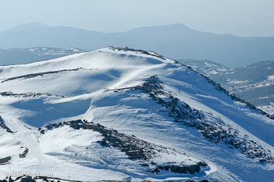 A mountain peak covered in deep snow with rocky ridges peeking through, more mountains are in the background