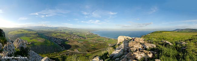 A panoramic view from a tall rocky cliff overlooking green hills on either side of a valley; the Sea of Galilee is visible, as well as some buildings and roads on the plain